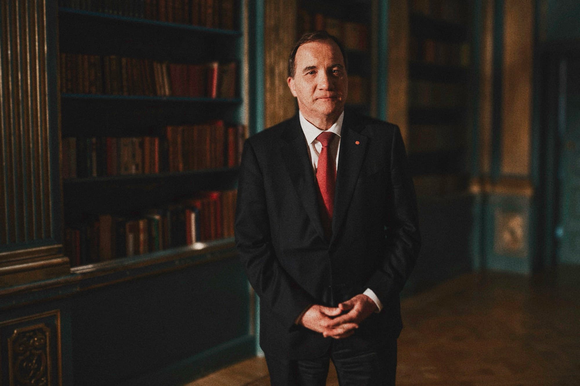 A man in a dark suit and red tie standing beside bookshelves
